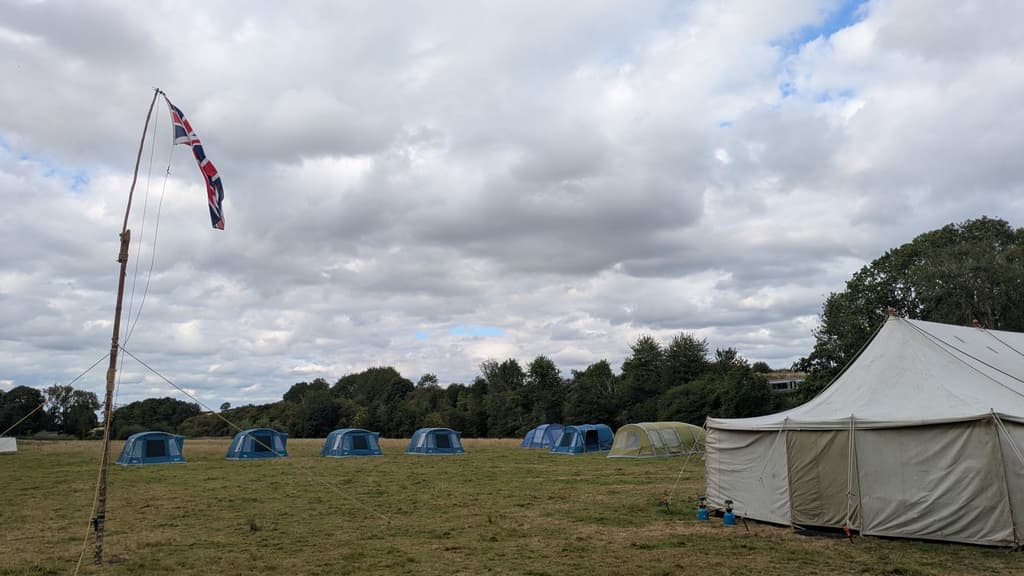Blue dome tents and a marquee in a field, with a Union flag flying from a lashed wooden pole